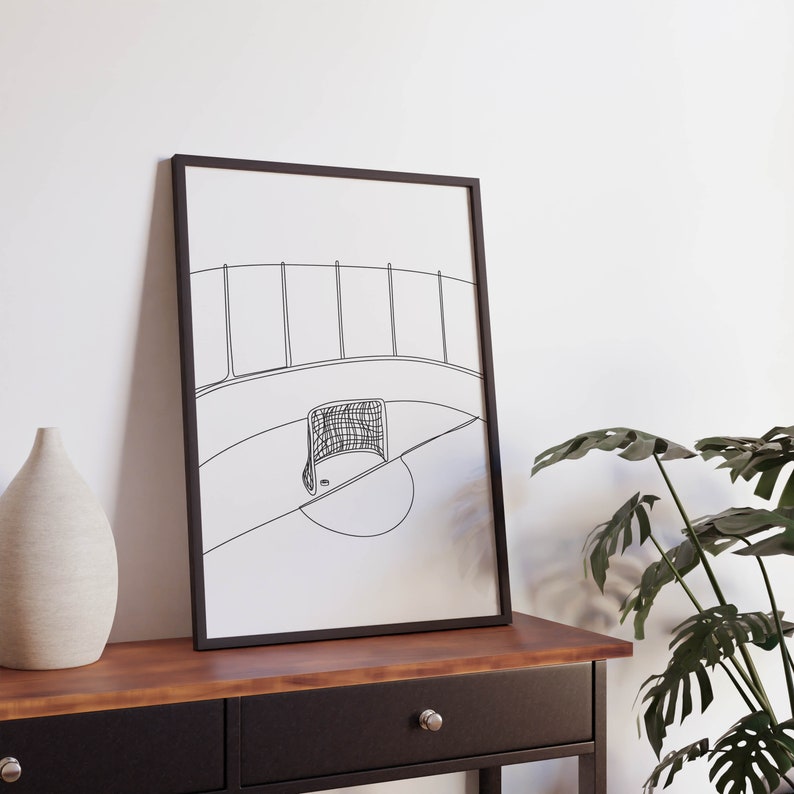 May include: Black and white line drawing of a hockey rink with a goal at center. The drawing is framed and hanging on a wall above a wooden table.