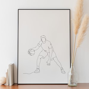 Peut inclure: Une impression encadrée noire d'un dessin au trait d'un joueur de basket-ball dribblant le ballon. L'impression est accrochée à un mur blanc au-dessus d'une commode en bois à deux tiroirs.
