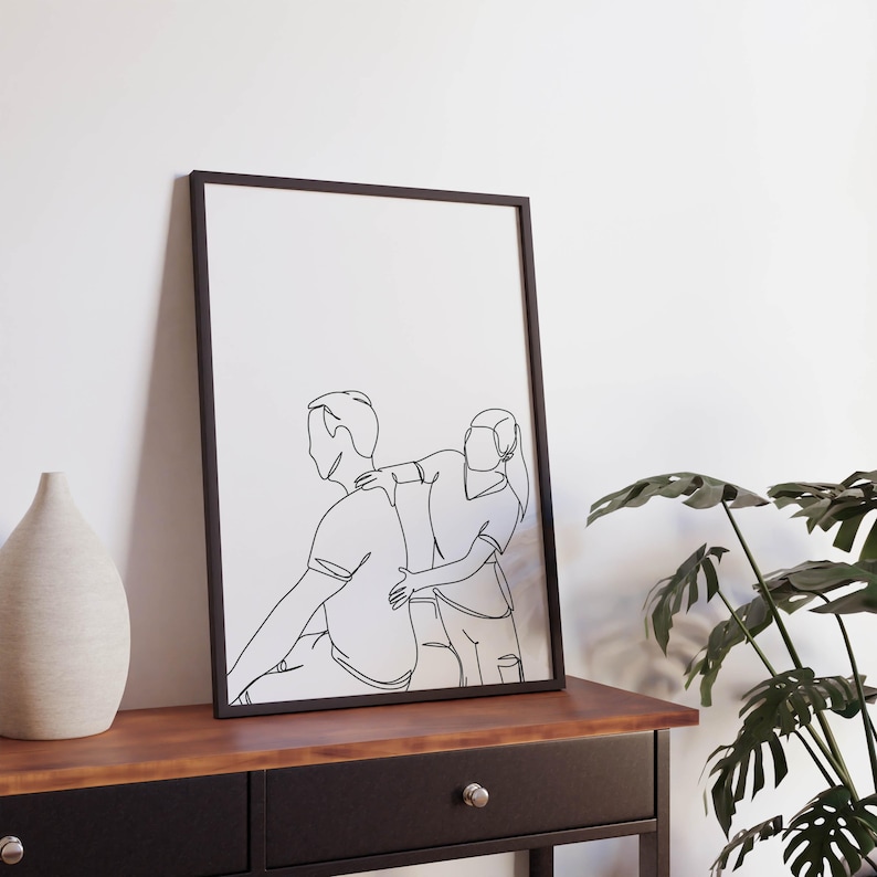 May include: A black framed print with a line drawing of a couple. The woman is standing behind the man, with her arm around his shoulder. The drawing is in a minimalist style.