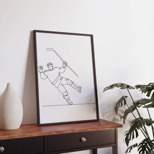 May include: A black and white line drawing of a hockey player skating and holding a hockey stick. The drawing is framed in a black frame and is hanging on a wall.