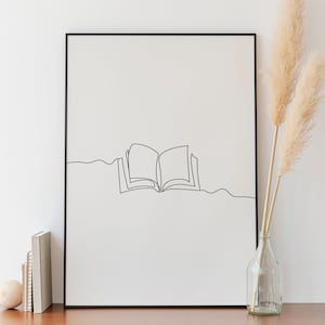 Könnte beinhalten: Ein schwarzer gerahmter Druck mit einer Strichzeichnung eines offenen Buches auf weißem Hintergrund. Der Druck hängt an einer weißen Wand über einer Kommode aus Holz mit zwei Schubladen. Eine Vase mit getrocknetem Pampasgras steht rechts neben der Kommode.