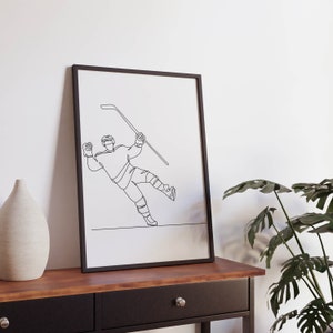 May include: Black and white line art print of a hockey player in a framed poster. The player is in mid-air, holding a hockey stick and about to shoot the puck.