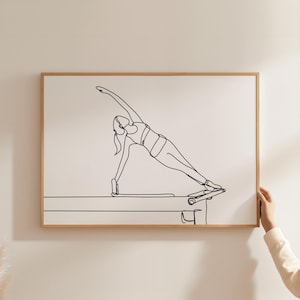 Puede incluir: Una impresión enmarcada que presenta un dibujo lineal minimalista de una persona realizando un ejercicio de Pilates en una máquina de reformer. La obra de arte es en negro sobre un fondo blanco, enmarcada en madera marrón claro.