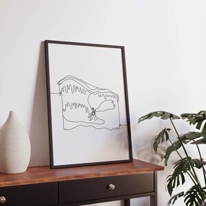 Puede incluir: Impresión artística enmarcada con un dibujo lineal minimalista de una persona escalando una montaña. La obra de arte es en negro sobre un fondo blanco. La impresión se exhibe sobre una mesa de madera oscura con una base negra, junto a un jarrón blanco y una planta verde.
