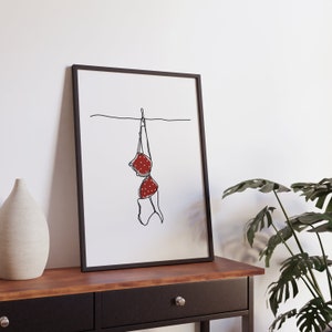 May include: Framed art print featuring a minimalist line drawing of a red polka-dot bra hanging on a clothesline. The artwork is set against a white background, with a black frame. The image is displayed on a wooden table with a decorative vase and a potted plant.