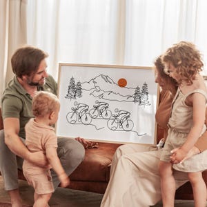 May include: A family of three looks at a framed black and white line drawing of cyclists riding through a mountain landscape. The drawing includes a mountain range, trees, and a sun.