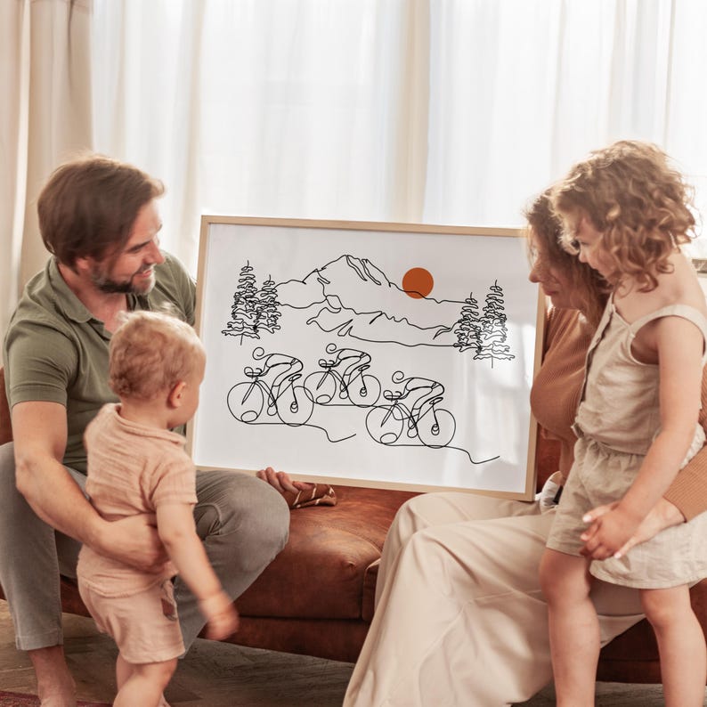 May include: A family of three looks at a framed black and white line drawing of cyclists riding through a mountain landscape. The drawing includes a mountain range, trees, and a sun.