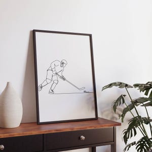 May include: Black and white line art print of a hockey player skating and shooting the puck. The print is framed in a black frame and is hanging on a white wall.