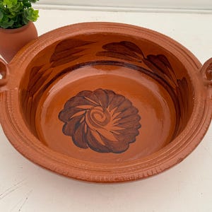 May include: A round, terracotta-colored clay bowl with two loop handles. The bowl features a dark brown floral design in the center and along the rim. A small terracotta pot with a green plant is in the background.