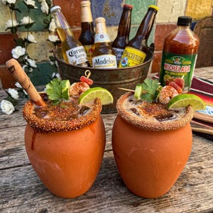 May include: Two terracotta clay cups filled with a dark liquid, rimmed with chili powder, and garnished with a lime wedge, cilantro, and a shrimp skewer. In the background, a metal bucket holds several beer bottles and a bottle of Michelada mix.