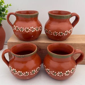 May include: Four terracotta mugs with handles. Each mug is a reddish-brown color with a green rim and a band of white and green floral-like designs around the middle. The mugs are sitting on a wooden surface.