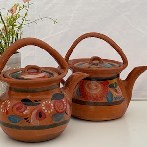 May include: Two terracotta teapots with matching lids and handles. The teapots are decorated with hand-painted floral designs in red, blue, and white. The teapots are sitting on a white surface.
