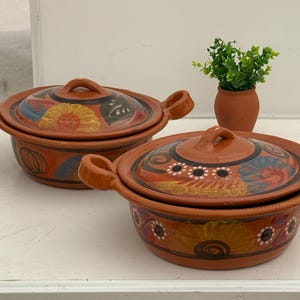 May include: Two terracotta cooking pots with lids. The pots have handles and are decorated with colorful floral patterns in orange, yellow, blue, and black. A small terracotta vase with a green plant is in the background.