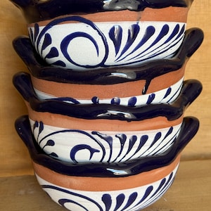 May include: A stack of four terracotta bowls with two handles each. The bowls feature a wavy rim glazed in dark blue, with a white band around the center decorated with hand-painted dark blue swirling floral patterns against the natural clay.