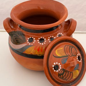 May include: A terracotta-colored ceramic pot with a matching lid. The pot features two handles and is decorated with black, white, yellow, and blue painted designs. The lid is partially removed, revealing the interior.