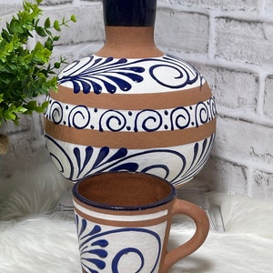 May include: A set of two terracotta pottery pieces, a large pitcher and a smaller mug, both with blue and white Talavera style designs. The pitcher has a wide, round body and a narrow neck, while the mug has a handle and a rounded shape.