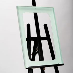 Könnte beinhalten: Ein hellgrüner Rahmen auf einem Staffelei. Der Rahmen ist aus Holz und hat eine rechteckige Form. Die Staffelei ist aus schwarzem Holz und hat ein traditionelles Design.