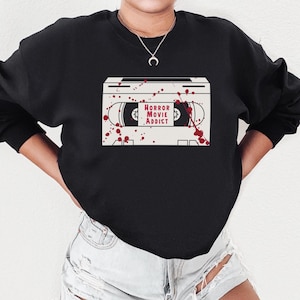 May include: Black sweatshirt featuring a graphic of a cassette tape with the words "HORROR MOVIE ADDICT" in red. The design includes red splatter details, creating a spooky aesthetic. The sweatshirt is a casual, comfortable style.