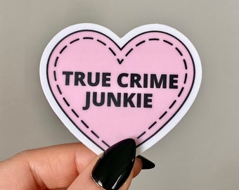 Może przedstawiać: Różowa naklejka w kształcie serca z białym obramowaniem i czarnymi szwami. Naklejka zawiera napis "TRUE CRIME JUNKIE" czarnym tekstem.