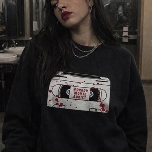 May include: Black sweatshirt with a white cassette tape graphic that reads "HORROR MOVIE ADDICT" with red blood splatter details. The sweatshirt is worn by a person with dark hair and red lipstick.