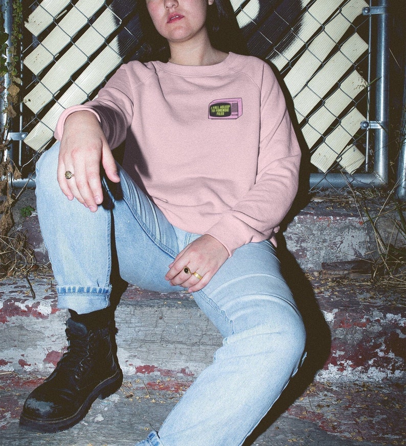 Op de afbeelding: Een lichtroze sweatshirt met een kleine retro tv-afbeelding met de tekst "I Will Sleep When I'm Famous". De sweatshirt wordt gecombineerd met een lichtblauwe jeans en zwarte laarzen.