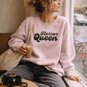 Może przedstawiać: Jasnor&oacute;żowa bluza z napisem "Horror Queen" w czarnym kolorze. Bluza jest noszona z ciemnoszarymi dżinsami. Aparat i okulary przeciwsłoneczne leżą na stole. Osoba trzyma nap&oacute;j kawowy.
