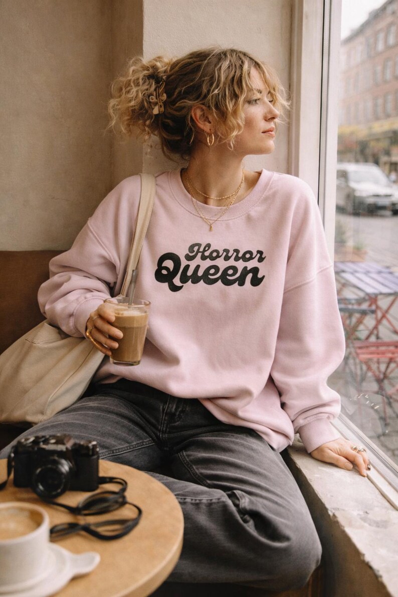 Może przedstawiać: Jasnor&oacute;żowa bluza z napisem "Horror Queen" w czarnym kolorze. Bluza jest noszona z ciemnoszarymi dżinsami. Aparat i okulary przeciwsłoneczne leżą na stole. Osoba trzyma nap&oacute;j kawowy.
