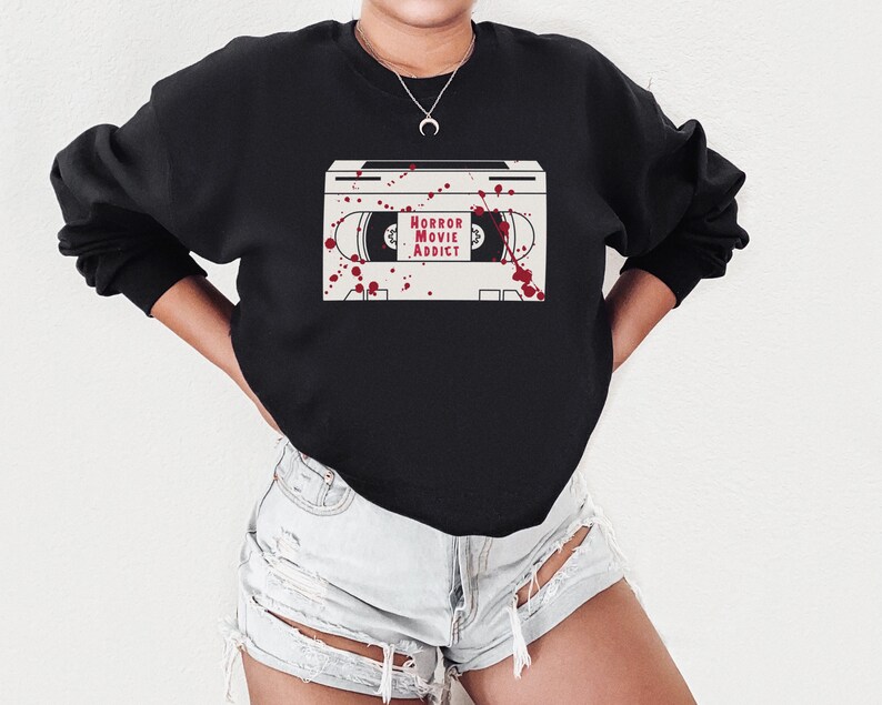 May include: Black sweatshirt featuring a graphic of a cassette tape with the words "HORROR MOVIE ADDICT" in red. The design includes red splatter details, creating a spooky aesthetic. The sweatshirt is a casual, comfortable style.