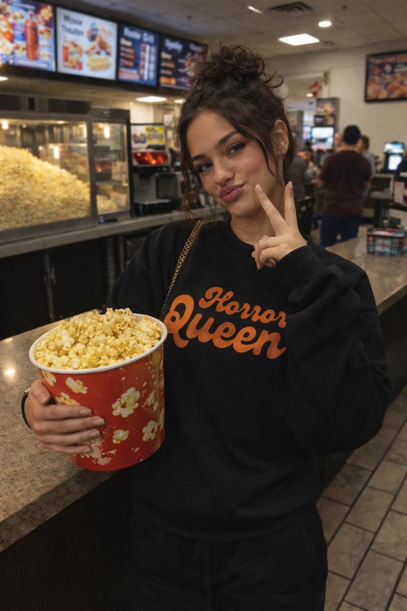 May include: A person wearing a black sweatshirt with the text "Horror Queen" in orange, holding a large red popcorn bucket with white floral designs. The bucket is filled with popcorn.