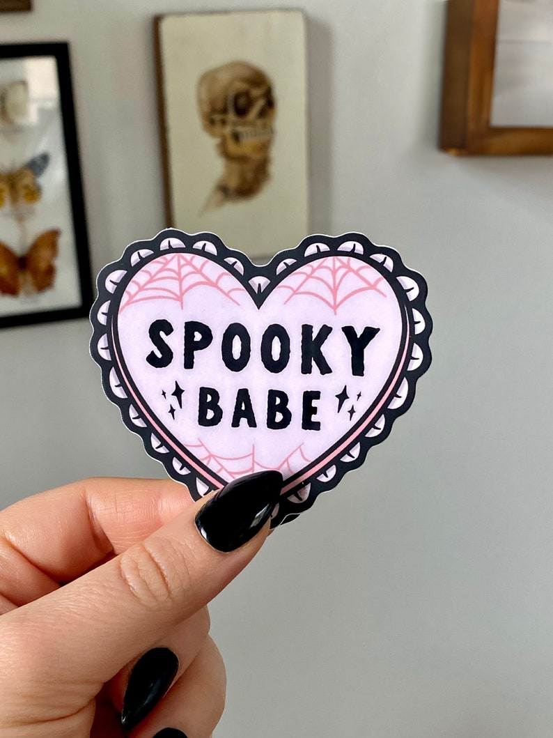May include: A pink heart-shaped sticker with a black lace border and a spiderweb design. The sticker says "SPOOKY BABE" in black letters with two small stars on either side of the word "BABE".