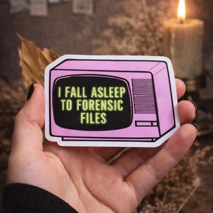 May include: A pink sticker featuring a retro television design with the text "I FALL ASLEEP TO FORENSIC FILES" in white. The sticker is held in a hand, with a lit candle in the background.