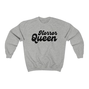 Może przedstawiać: Jasnoszara bluza z napisem "Horror Queen" w czarnym, retro kroju pisma. Bluza ma okrągły dekolt i długie rękawy. Tekst jest wyśrodkowany na klatce piersiowej.