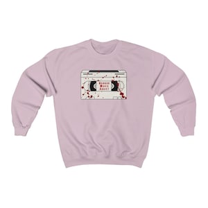 May include: A light pink sweatshirt featuring a graphic of a white VHS tape with the words "HORROR MOVIE ADDICT" in red. Red blood splatter details are around the tape. The sweatshirt has long sleeves and a crew neck.