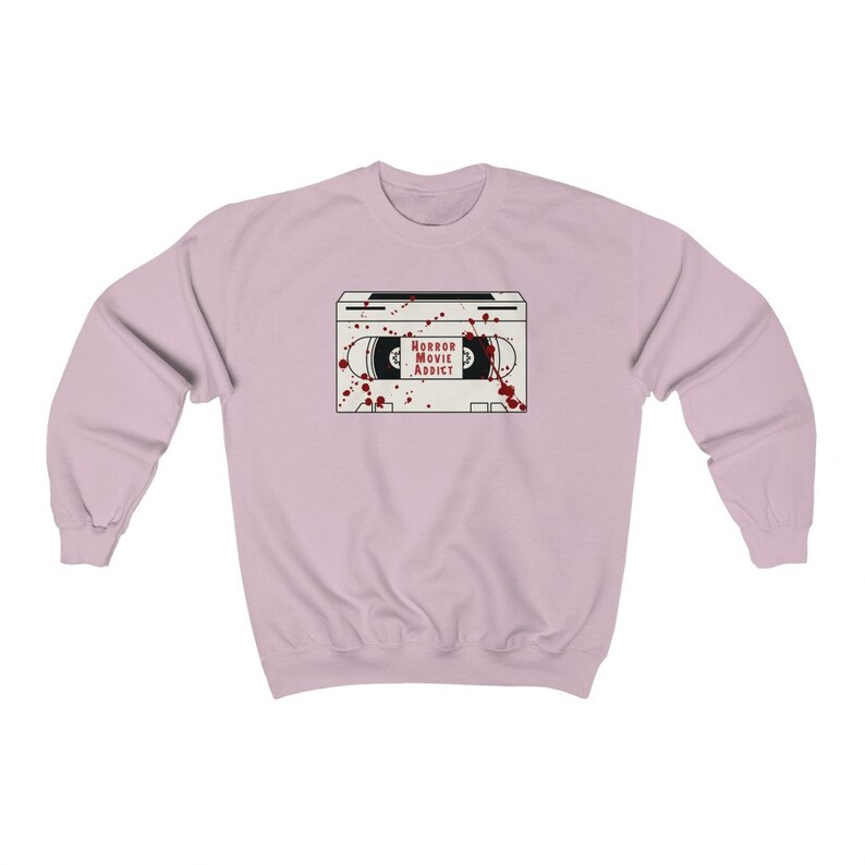 May include: A light pink sweatshirt featuring a graphic of a white VHS tape with the words "HORROR MOVIE ADDICT" in red. Red blood splatter details are around the tape. The sweatshirt has long sleeves and a crew neck.