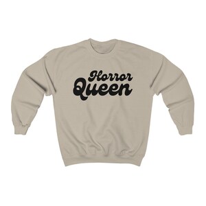 Może przedstawiać: Beżowa bluza z napisem "Horror Queen" w czarnym, retro kroju pisma. Odzież ma okrągły dekolt i długie rękawy. Tekst jest wyśrodkowany na piersi.