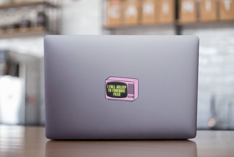 May include: A sticker featuring a pink retro television with the text "I FALL ASLEEP TO FORENSIC FILES" in a green font. The sticker is placed on the back of a gray laptop.
