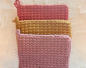Puede incluir: Tres agarraderas de olla de ganchillo en rosa, amarillo y rojo. Las agarraderas están apiladas una encima de la otra.