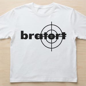 Könnte beinhalten: Ein weißes T-Shirt mit kurzen Ärmeln und einem schwarzen Grafikdruck auf der Brust. Das Design besteht aus dem Wort "bratort" in einer fetten serifenlosen Schrift, wobei ein schwarzes Fadenkreuz-Symbol über den letzten Buchstaben liegt.
