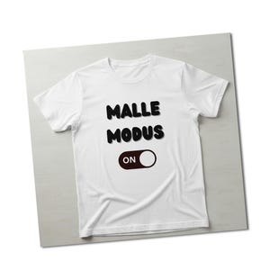 Könnte beinhalten: Weißes T-Shirt mit den Worten "MALLE MODUS" in schwarzen, fetten Buchstaben. Darunter befindet sich ein brauner und weißer Kippschalter mit dem Wort "ON". Das Shirt wird auf einer hellgrauen Oberfläche präsentiert.