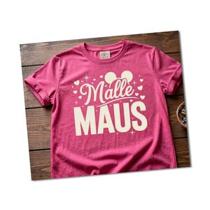 Könnte beinhalten: Ein magentafarbenes T-Shirt mit hochgekrempelten Ärmeln. Das Shirt zeigt den Text "Malle MAUS" in Weiß, mit Mickey Mouse Ohren und Herz-Akzenten. Das T-Shirt liegt auf einer Holzoberfläche.