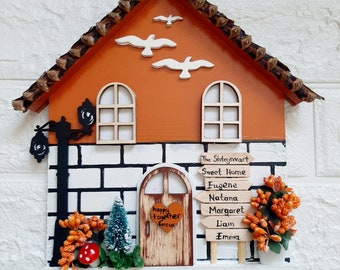 Puede incluir: Un portallaves de madera con forma de casa con una pared de ladrillo, un árbol pequeño y una puerta con las palabras "happy together forever". La casa tiene un techo marrón con un borde blanco y una base blanca. Hay tres ganchos en la parte inferior para colgar las llaves. La casa tiene un letrero que dice "The Shtenyvwart Sweet Home Eugene Natana Margaret Liam Emma".