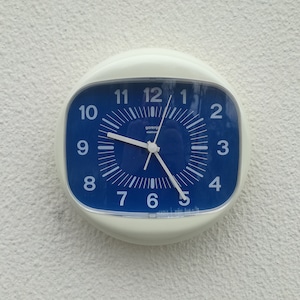 Könnte beinhalten: Eine weiße und blaue Wanduhr mit einem quadratischen Zifferblatt und schwarzen Zeigern. Das Zifferblatt der Uhr ist blau mit weißen Zahlen und einem weißen Rand. Die Uhr hängt an einer weißen Wand.