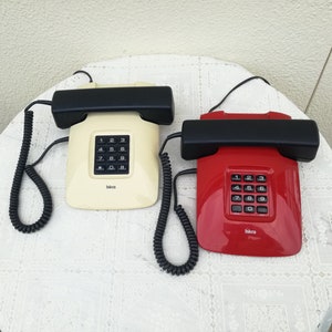 Op de afbeelding: Twee vintage draaischijftelefoons, een wit en een rood, met zwarte hoornen en opgerolde snoeren. Beide telefoons hebben een zwart toetsenbord met de merknaam "takora" onderaan.