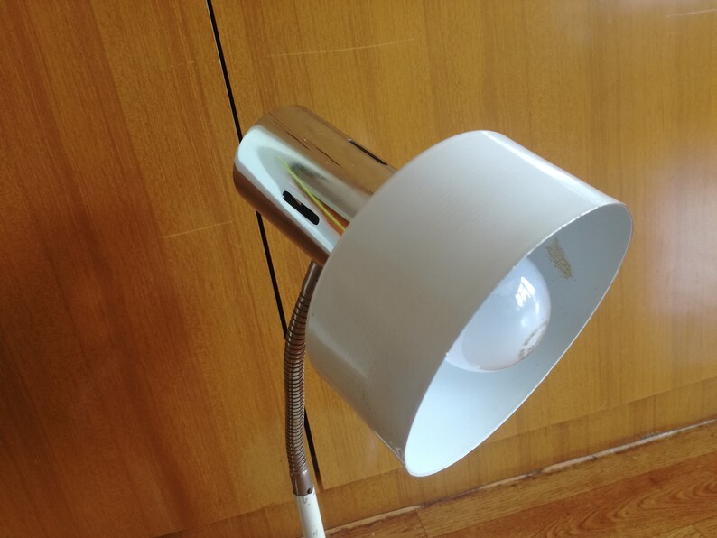 May include: A white and chrome desk lamp with a flexible arm and a white shade. The lamp is turned on and the light bulb is visible.