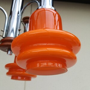 Puede incluir: Primer plano de una l&aacute;mpara de ara&ntilde;a moderna de mediados de siglo en cromo y naranja con m&uacute;ltiples luces. Las pantallas de vidrio naranja tienen un dise&ntilde;o en capas y redondeado.