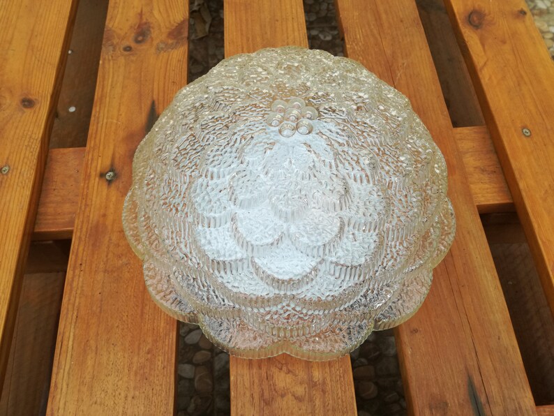 May include: A clear glass ceiling light fixture with a textured, flower-like design. The light fixture has a rounded shape and a scalloped edge.