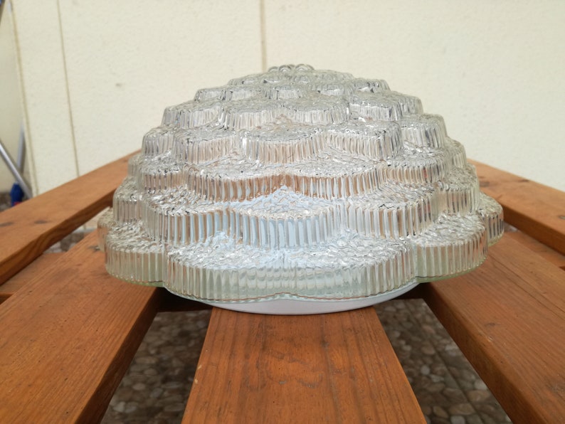 May include: A clear glass ceiling light fixture with a textured, flower-like design. The light fixture is made of multiple layers of glass, creating a unique and intricate pattern.