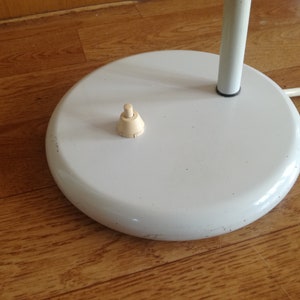 May include: A white lamp base with a round, flat top and a white cord. The base has a small, off-white switch on the top.