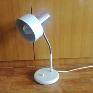 May include: A white and chrome desk lamp with a flexible arm and a white shade. The lamp is on a wooden floor.