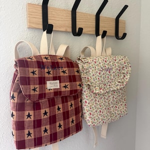May include: Two backpacks hanging on a wooden rack with four black hooks. The left backpack features a red and dark red plaid pattern with black stars and the name Walker. The right backpack has a floral print with the name Josie embroidered.